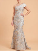 A woman models a floor-length one-shoulder evening gown with a fitted silhouette and flared mermaid hem. The dress features an intricate metallic floral pattern in silver and gold tones.
