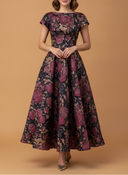 A woman modeling a floor-length A-line dress with a floral jacquard pattern in deep burgundy, purple, and beige on a dark navy base, set against a solid warm tan studio background.